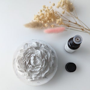 Puede incluir: Difusor de cerámica blanco con un diseño de flor en relieve. El difusor es redondo y tiene una pequeña abertura en la parte superior. Una pequeña botella de aceite esencial está al lado del difusor.
