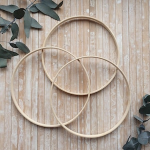 May include: Three natural wooden embroidery hoops of different sizes are arranged on a light wood-grain surface, with eucalyptus leaves in the corners. The hoops overlap, creating a pattern. The wood has a light, unfinished appearance.