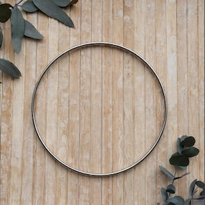 May include: A circular silver metal hoop, likely a wreath frame, sits on a light brown wooden surface. Eucalyptus leaves are arranged around the hoop, adding greenery. The hoop's shape contrasts with the vertical wood grain.