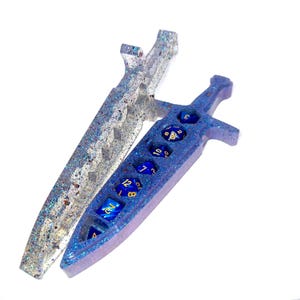 May include: Two resin dice holders shaped like swords. One is silver with glitter, the other is blue with glitter and holds blue dice. The dice have gold numbers. The sword-shaped holders are for tabletop gaming.