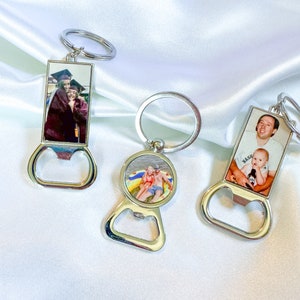 Puede incluir: Tres abridores de botellas de plata con insertos de fotos personalizadas. El primer llavero presenta una foto de dos personas con atuendos de graduaci&oacute;n. El segundo llavero presenta una foto de dos personas en un flotador en una piscina. El tercer llavero presenta una foto de un hombre y un ni&ntilde;o. El llavero tiene la palabra "Nash" impresa.