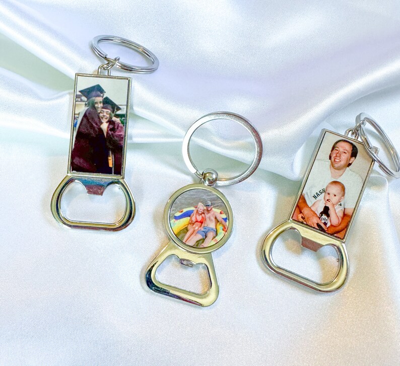 Puede incluir: Tres abridores de botellas de plata con insertos de fotos personalizadas. El primer llavero presenta una foto de dos personas con atuendos de graduaci&oacute;n. El segundo llavero presenta una foto de dos personas en un flotador en una piscina. El tercer llavero presenta una foto de un hombre y un ni&ntilde;o. El llavero tiene la palabra "Nash" impresa.