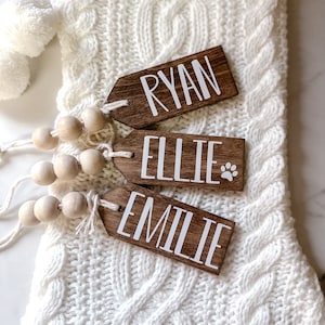 May include: Three wooden gift tags with white lettering, each with a different name: Ryan, Ellie, and Emilie. The tags are attached to a white knitted stocking with wooden beads.