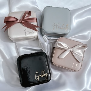 May include: Four small square jewelry boxes with personalized names. The boxes are in different colors: light gray, black, beige, and white. Each box has a satin ribbon tied around it.
