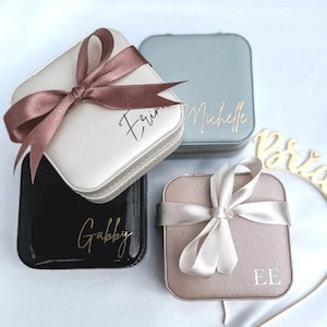 May include: Four square jewelry boxes in cream, black, dusty rose, and gray, each with a satin ribbon bow. The boxes have gold lettering with names and initials. A gold "Bride" decoration is in the background.