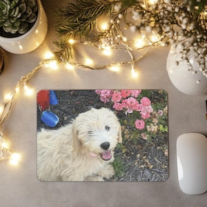 May include: Rectangular mousepad featuring a light-colored dog with a pink collar, positioned in front of pink flowers and blue and red objects. The mousepad is on a gray surface, with a white mouse to the right.