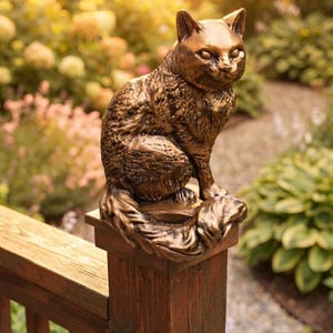 May include: A bronze-colored cat statue in a seated position with its tail wrapped around its body. The sculpture features detailed fur texture and pointed ears, positioned atop a square wooden post. The finish has a metallic sheen.