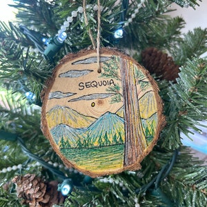 May include: A wooden Christmas ornament with a hand-painted scene of a forest with mountains and a tall tree. The ornament is painted in shades of green, brown, and blue. The word "SEQUOIA" is written on the ornament.