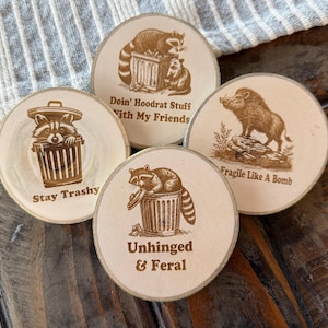 May include: Four wooden coasters with different designs and sayings. The coasters feature a raccoon, a wild boar, and a trash can. The sayings on the coasters are "Stay Trashy", "Doin' Hoodrat Stuff With My Friends", "Fragile Like A Bomb", and "Unhinged & Feral".