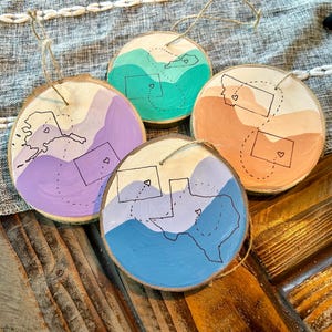 May include: Four wooden ornaments painted with different color gradients and outlines of states with hearts in the middle. The states are Alaska, Kentucky, Montana, and Texas.