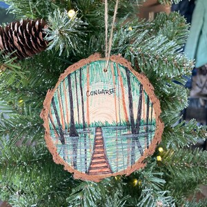May include: A wooden Christmas ornament with a hand-painted scene of a forest with a wooden bridge over a lake. The ornament is decorated with green, brown, and blue paint and has the word "Congaree" written on it.
