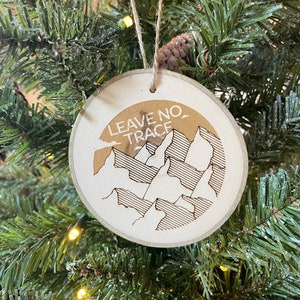 May include: A wooden Christmas ornament with a brown background and white text that reads "Leave No Trace" with a mountain range graphic.