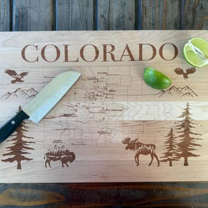 May include: A wooden cutting board with the word "COLORADO" engraved on it. The board features a map of Colorado with etched illustrations of wildlife and landmarks. A knife and lime halves are on the board.