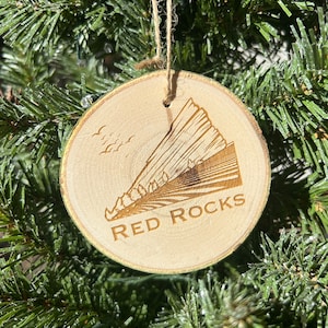 May include: A wooden Christmas ornament with a laser-etched design of Red Rocks Amphitheatre in Colorado. The ornament is hanging on a green Christmas tree.