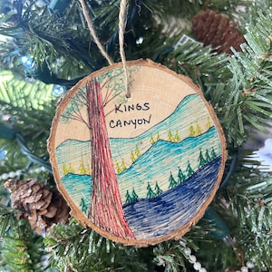 May include: A wooden Christmas ornament with a hand-painted scene of a redwood tree and mountains. The ornament is painted in shades of blue, green, and red. The text "Kings Canyon" is written on the ornament.