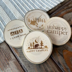 May include: Four wooden coasters with a camping theme. The coasters feature different designs, including a tent, trees, a bear, a moon, and the text "Apparently, I camp now", "Unhappy Camper", and "Happy Campers".