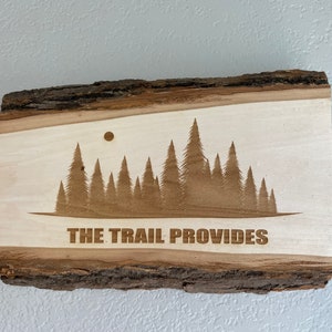 May include: A wooden sign with a natural bark edge, featuring a laser-etched design of a forest silhouette with a moon above. The text "THE TRAIL PROVIDES" is also laser-etched on the sign.
