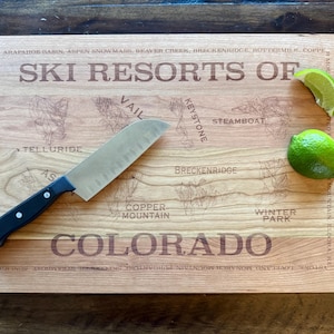 May include: Wooden cutting board with the text "SKI RESORTS OF COLORADO" and illustrations of ski resorts. A black-handled knife and two lime wedges are on the board. The board is rectangular and made of light-colored wood.