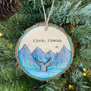 May include: A wooden ornament with a blue and white painted scene of mountains and a whale tail. The text "KENAI FJORDS" is written on the ornament.