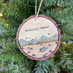 May include: A wooden Christmas ornament with a purple edge, featuring a hand-drawn illustration of a petrified forest landscape with mountains, a sun, and a log. The ornament is labeled "Petrified Forest".