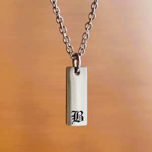 May include: A silver chain necklace with a rectangular pendant featuring a black letter 'B' in an old English font.