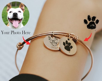 May include: A gold bangle bracelet with two charms. One charm has a black and white photo of a dog's face with the name "Max" engraved below. The other charm has a black paw print.