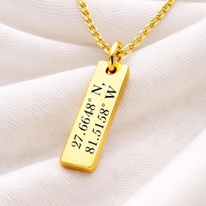 May include: A gold-colored necklace with a rectangular pendant. The pendant has the coordinates 27.6648° N, 81.5158° W engraved in black. The chain is a delicate link style, and the background is a white fabric.