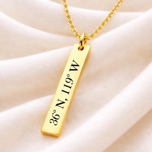 May include: A gold-colored necklace with a rectangular pendant. The pendant is engraved with the coordinates "36° N, 119° W" in black lettering. The necklace chain is also gold-colored and hangs against a white fabric background.