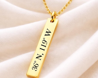 May include: A gold-colored necklace with a rectangular pendant. The pendant is engraved with the coordinates "36° N, 119° W" in black lettering. The necklace chain is also gold-colored and hangs against a white fabric background.