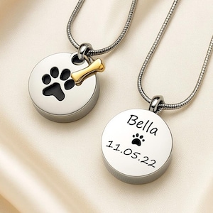 May include: Two silver pet memorial necklaces with paw print designs. One pendant has a black paw print and a gold bone charm. The other pendant is engraved with the name "Bella", a paw print, and the date 11.05.22.