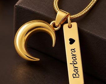 May include: A gold-toned necklace featuring a crescent moon pendant and a rectangular vertical bar charm. The bar charm is engraved with a small black heart symbol above the name Barbara in a cursive script font, suspended on a gold snake chain.