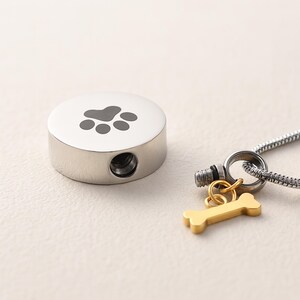 May include: A silver-colored pet urn with a paw print design on top. A small gold-colored bone charm hangs from a silver chain. The urn has a small opening on the side.