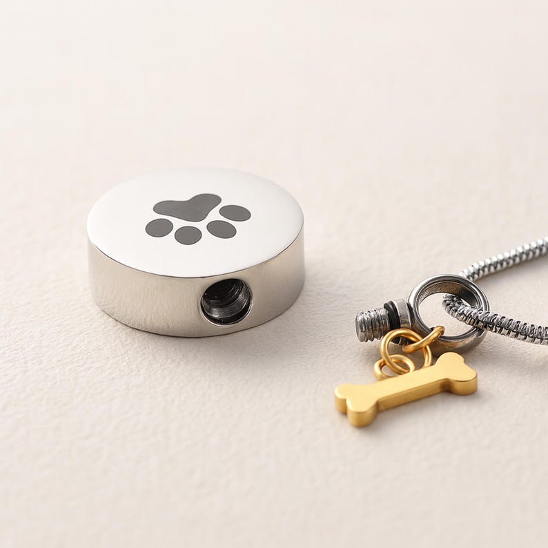 May include: A silver-colored pet urn with a paw print design on top. A small gold-colored bone charm hangs from a silver chain. The urn has a small opening on the side.