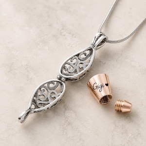 Pu&ograve; includere: Una collana con ciondolo color argento caratterizzata da due gabbie in filigrana a forma di goccia collegate. Accanto al ciondolo si trova un tappo cilindrico color oro rosa inciso con il nome Gigi e un piccolo cuore, con una vite filettata.