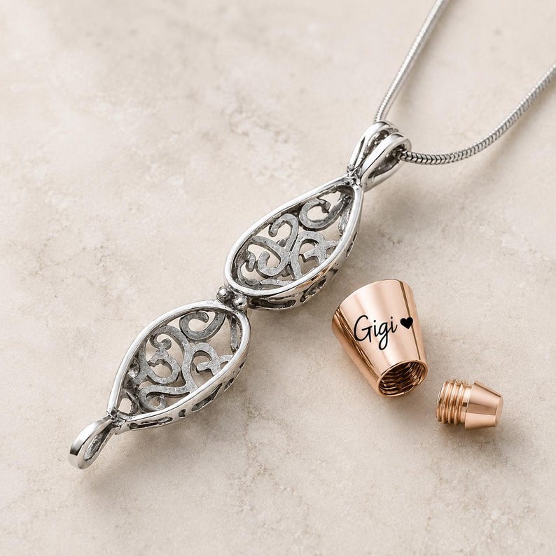 Pu&ograve; includere: Una collana con ciondolo color argento caratterizzata da due gabbie in filigrana a forma di goccia collegate. Accanto al ciondolo si trova un tappo cilindrico color oro rosa inciso con il nome Gigi e un piccolo cuore, con una vite filettata.