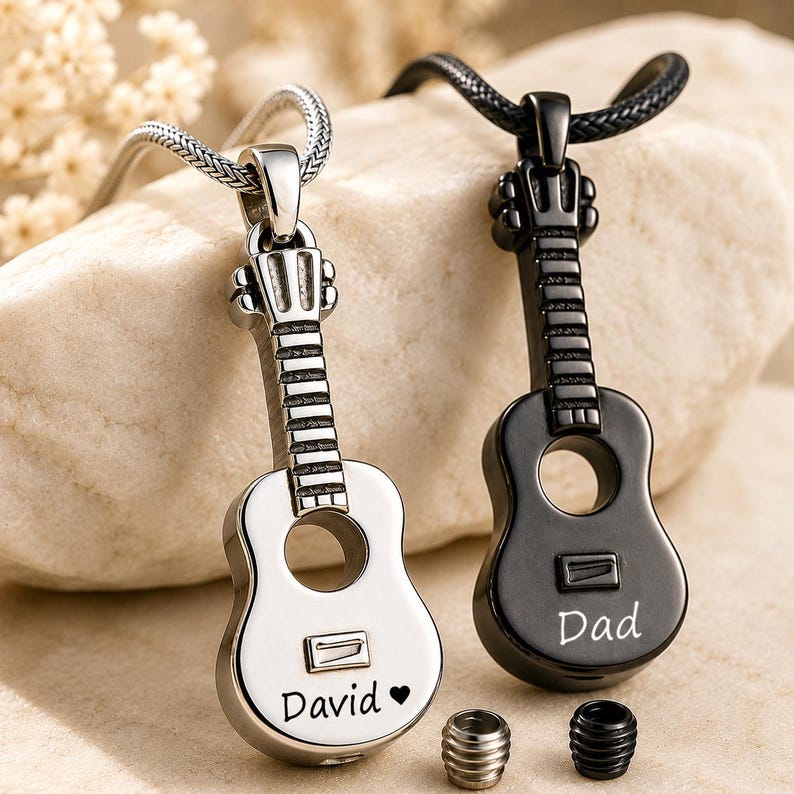 K&ouml;nnte beinhalten: Zwei gitarrenf&ouml;rmige Anh&auml;nger, einer aus poliertem silberfarbenem Metall mit der Gravur David und einem Herz, der andere aus mattem schwarzem Metall mit der Gravur Dad. Jeder Anh&auml;nger verf&uuml;gt &uuml;ber eine kleine Gewindeschraubkappe an der Basis.