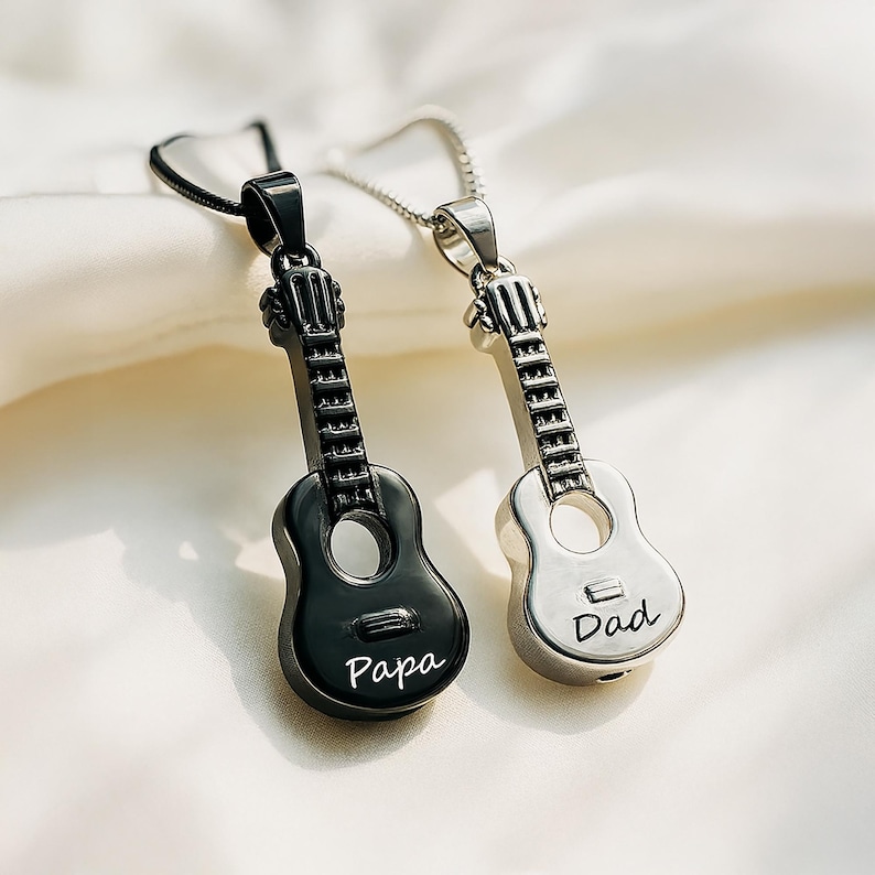K&ouml;nnte beinhalten: Zwei gitarrenf&ouml;rmige Anh&auml;nger an Ketten. Ein Anh&auml;nger ist schwarz und tr&auml;gt das Wort Papa auf dem Korpus, der andere ist silberfarben und tr&auml;gt das Wort Dad auf dem Korpus. Beide zeigen einen Hals, eine Kopfplatte und ein Schallloch.