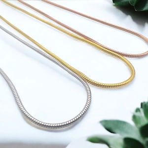 May include: Three snake chains in silver, gold, and rose gold. The chains are laid out on a white surface.
