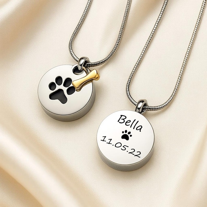 May include: Two silver pet memorial necklaces with paw print designs. One pendant has a black paw print and a gold bone charm. The other pendant is engraved with the name "Bella", a paw print, and the date 11.05.22.