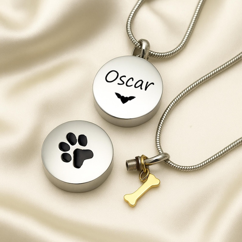 May include: Two silver-colored pet memorial necklaces. One pendant is round with the name "Oscar" and a small heart design. The other pendant is round with a black paw print. A gold-colored bone charm hangs from one necklace.