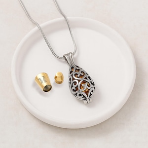 Pu&ograve; includere: Una collana con ciondolo a forma di goccia color argento con un intricato design in filigrana e un interno color oro. Accanto al ciondolo si trova un piccolo tappo cilindrico color oro staccato con il testo inciso "Miss you" sul lato.