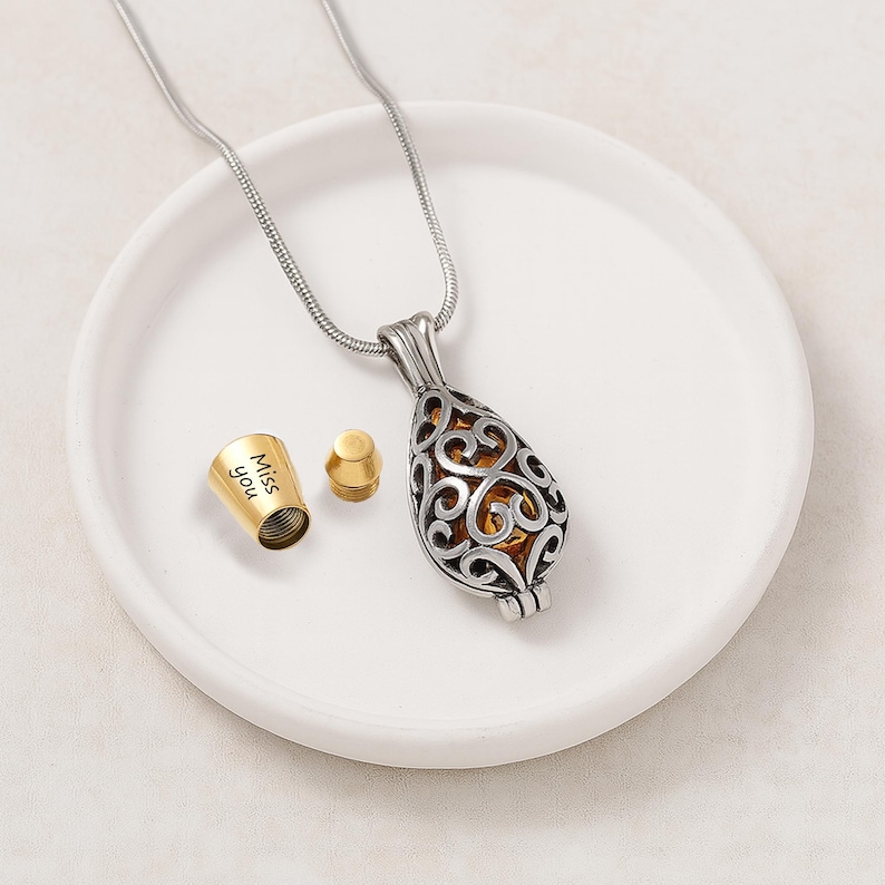 Pu&ograve; includere: Una collana con ciondolo a forma di goccia color argento con un intricato design in filigrana e un interno color oro. Accanto al ciondolo si trova un piccolo tappo cilindrico color oro staccato con il testo inciso "Miss you" sul lato.