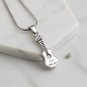 K&ouml;nnte beinhalten: Ein silberfarbener Metallanh&auml;nger in Form einer akustischen Gitarre, der an einer passenden Schlangenkette h&auml;ngt. Der Gitarrenk&ouml;rper tr&auml;gt die Gravur 'Miss you' und enth&auml;lt ein kleines, abnehmbares Gewindeschraubteil zum Verschlie&szlig;en.