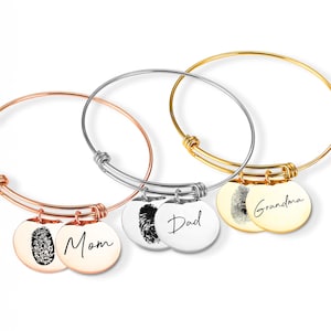 May include: Three bangle bracelets in rose gold, silver, and gold. Each bracelet has a round charm with a fingerprint and a name: Mom, Dad, and Grandma. The bracelets are on a white background.