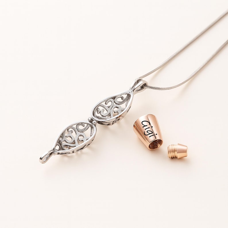 Pu&ograve; includere: Una collana color argento con un ciondolo in filigrana traforata a forma di goccia, accompagnata da un tappo cilindrico color oro rosa inciso con il nome Gigi e un piccolo componente metallico a vite coordinato per la chiusura.