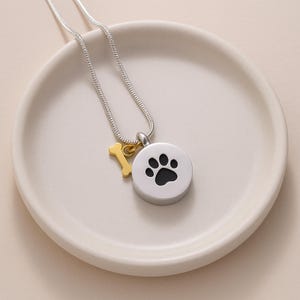May include: A silver-colored necklace with a round pendant featuring a black paw print. A small gold-colored bone charm hangs next to the pendant. The necklace is displayed on a white, shallow dish.