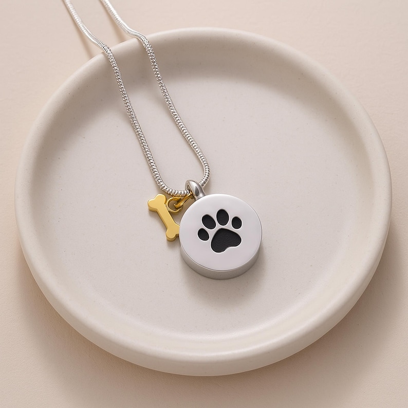May include: A silver-colored necklace with a round pendant featuring a black paw print. A small gold-colored bone charm hangs next to the pendant. The necklace is displayed on a white, shallow dish.
