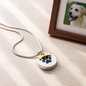 May include: A silver necklace with a round, white pendant featuring a black paw print and a gold-colored bone charm. A framed photo of a dog is in the background. The pendant is a memorial item.