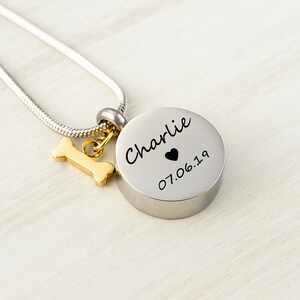 May include: A silver-colored pendant necklace with a round, engraved charm. The charm has the name "Charlie", a heart, and the date "07.06.19". A gold-colored bone charm hangs next to the round charm.