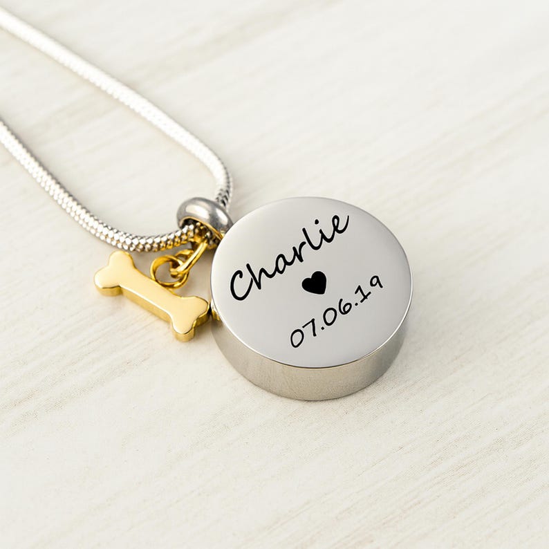 May include: A silver-colored pendant necklace with a round, engraved charm. The charm has the name "Charlie", a heart, and the date "07.06.19". A gold-colored bone charm hangs next to the round charm.